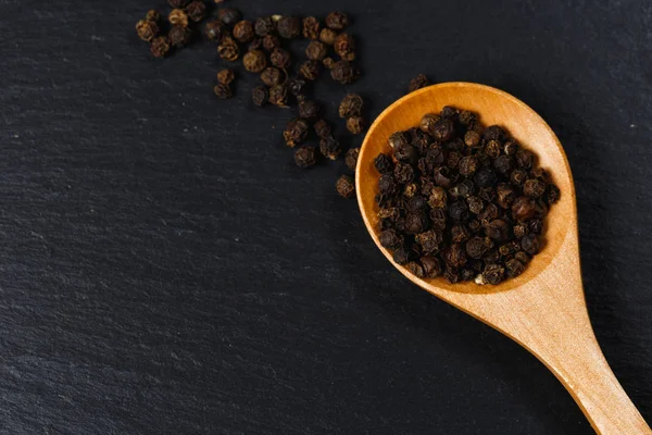 Black pepper spice in wooden spoon. Flat lay of black pepper spice on black stone. Black pepper spice, top view. Organic food, healthy lifestyle. Space for text