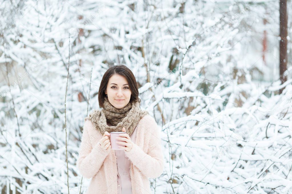 girl in a pink sweater drinking coffee under the falling snow in the winter forest