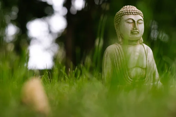 Piccola statua di Buddha bianco in una posa di meditazione con erba verde in primo piano e su sfondo naturale sfocato luminoso. Simbolo religioso del buddismo. Focus selettivo — Foto Stock