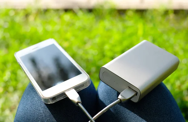 Beyaz akıllı telefon yeşil çim arka plan üzerinde diz üzerinde güç bankası tarafından şarj. Seyahat ederken veya kamp yaparken bağlı kalın.