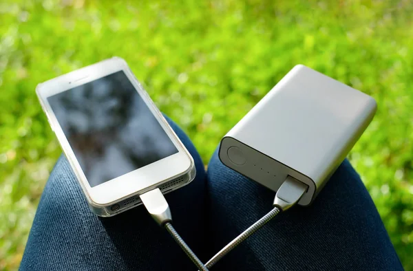 Bílý smartphone nabitý z elektrické energie na kolenou na pozadí zelené trávy. Zůstaňte při cestování nebo kempování připojeni. — Stock fotografie