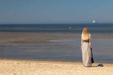 Ust-Narva, Ida-Viru County, Estonya, Ida-Viru County, Estonya, 23 Haziran 2020. Kumsalda, güneşli bir günde, uzun gri elbiseli bir kız denize bakıyor.