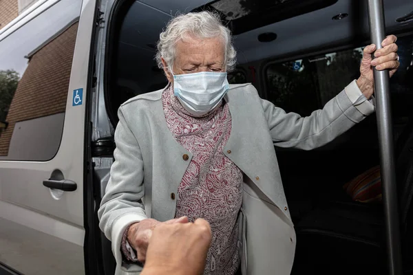 Tekerlekli sandalyeli, tıbbi maskeli yaşlı bir kadın engelli birini arabaya bindiriyor..