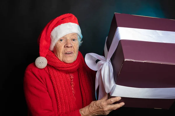 새 년의 모자를 쓴 명랑 한 노인, 그녀의 손에 큰 선물. 스튜디오 사진. 어두운 배경. 크리스마스 축하 해. — 스톡 사진