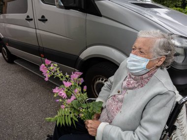 Elinde çiçekle tıbbi maske takan yaşlı bir kadın. Tekerlekli sandalyedeki engelli birinin hastaları arabayla taşıması fikri.