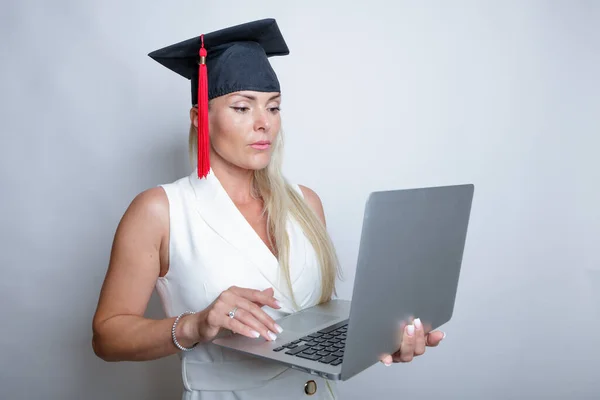 Belle casquette de graduation blonde tient un ordinateur portable dans sa main, fond lumineux studio. Concept, éducation, apprentissage à distance, en ligne — Photo