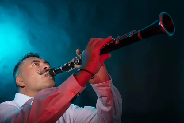 A man with a long, imposing mustache plays the clarinet. Studio, dark background, blue illumination — Stock Photo, Image