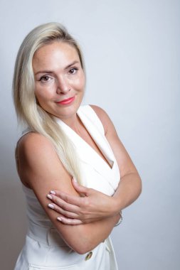 Beyaz takım elbiseli güzel sarışın kamera önünde poz veriyor, stüdyo ışığı arka planda.