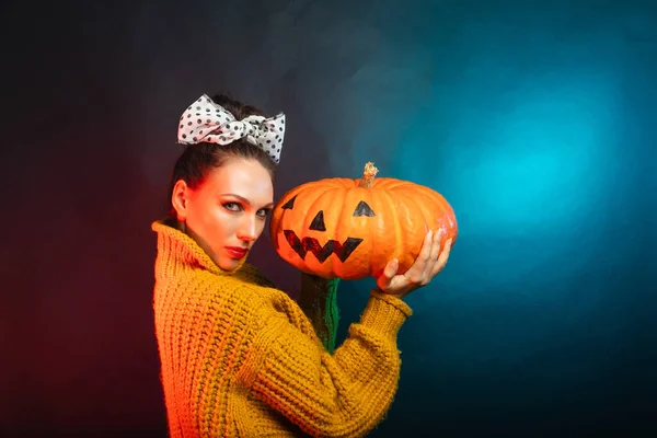 A brunette with a polka-dot bow and a yellow sweater holding a pumpkin. In the studio dark background Halloween concept. There is a place for text — Stock Photo, Image