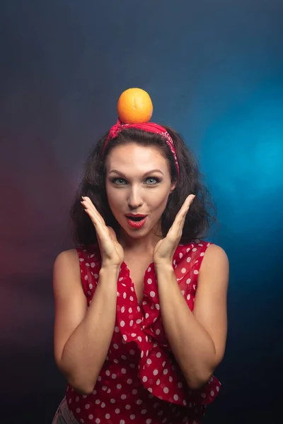 Brunette with an orange. Studio, dark background She holds a fruit on her head.