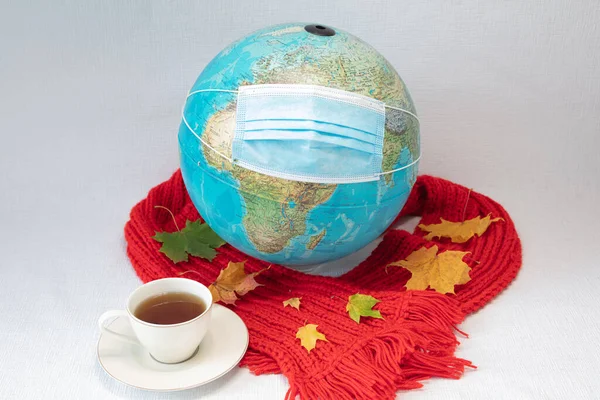The globe is wearing a medical mask and red warm scarf with maple leaves and a cup of tea. white background. concept, our world is infected with coronavirus. Autumn, stay at home in quarantine
