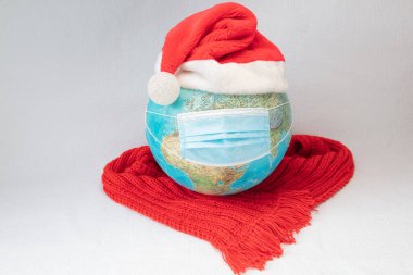 Dünya tıbbi bir maske takıyor Noel şapkası ve kırmızı sıcak bir eşarp. Beyaz arka plan. Kavram, dünyamız koronavirüs tarafından enfekte edilmiştir. Noel, yeni yıl evde karantina altında