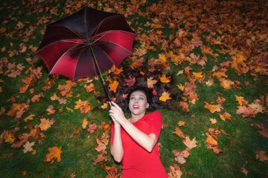 Uzun saçlı esmer bir kadının portresi. Kırmızı bir elbiseyle yeşil çimenlerin üzerine uzanır ve saçındaki akçaağaç yaprakları elinde bir şemsiye tutar. Sonbahar konsepti ..