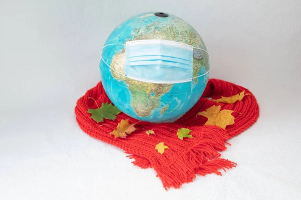 A medical mask and a red warm scarf with maple leaves are wearing on the globe. White background. concept, our world is infected with coronavirus. Autumn, stay at home in quarantine