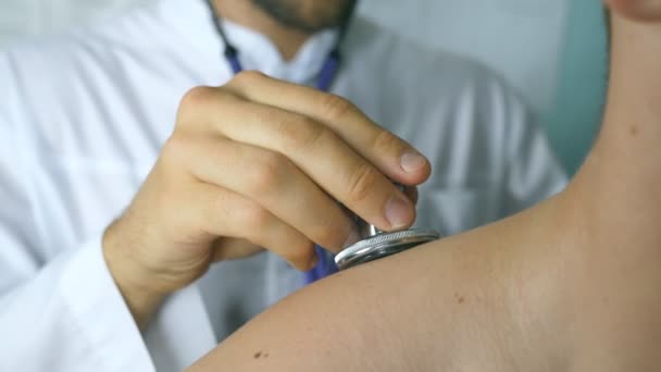 Doctor examinando a un paciente joven con estetoscopio. Médico revisando el pecho de un tipo en su oficina en el hospital. Trabajador médico escuchando el latido del corazón del hombre. Primer plano Vista trasera trasera — Vídeos de Stock