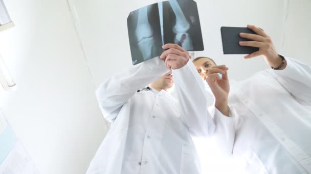 Mężczyźni medycy korzystający z tabletu podczas konsultacji ze sobą na temat rentgenowskiego obrazu pacjenta. Pracownicy medyczni w szpitalu badają odciski rentgenowskie. Dwóch kaukaskich lekarzy ogląda zdjęcie i dyskutuje o tym — Wideo stockowe