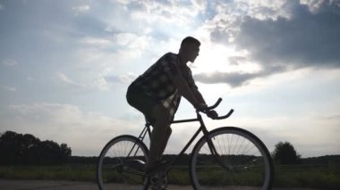 Genç adam arka plan güzel günbatımı gökyüzüne vintage bisiklet sürme silüeti. Sportif adam ülkede yol Bisiklete binme. Erkek bisikletçi sabit binicilik bisiklet dişli. Sağlıklı yaşam yavaş hareket