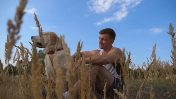 Jonge man zittend op groen gras op de weide en zijn hond aaien. Mannelijke eigenaar tijd samen met zijn labrador / golden retriever op aard. Close-up van Slow motion — Stockvideo