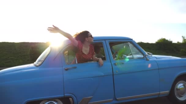 Chica feliz en gafas de sol que se inclina por la ventana del coche retro y disfrutar del viaje. Mujer joven mirando por la ventana de mover viejo auto en el día soleado. Concepto de viaje y libertad. Cámara lenta Primer plano — Vídeos de Stock