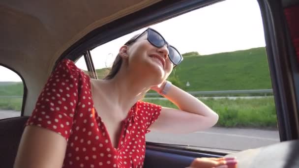 Gelukkig meisje in een zonnebril leunend uit het retro autoraam en genietend van de reis. Jonge vrouw kijkt uit het raam van het verplaatsen van oude vintage auto op zonnige dag. Reizen en vrijheid concept. Langzame beweging Close-up — Stockvideo