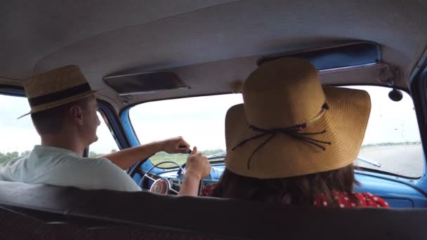 年轻夫妇的帽子骑在老式汽车在夏季旅行。男子和妇女坐在旧复古汽车的前排座位, 并给予对方五。道路旅行概念。慢动作后向后视图 — 图库视频影像