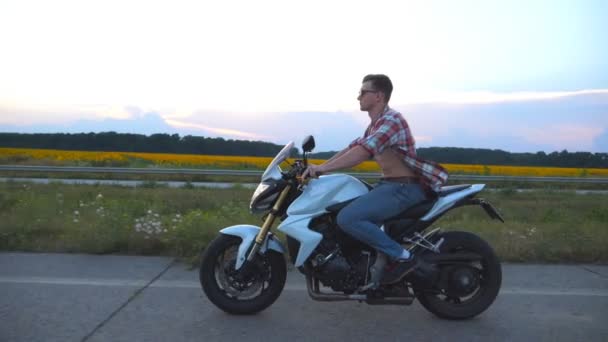 Young man riding on a modern sport motorbike. Handsome motorcyclist in a shirt and glasses driving his motorcycle on country road with sunflowers field at background. Slow motion Side view Close up — Stock Video