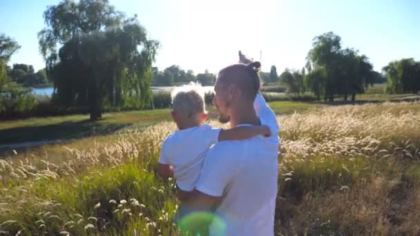 Молодий тато тримає дитину на лузі. Батько і син вказують на сонце на полі. Щаслива сім'я проводить час разом. Літній пейзаж на фоні. Крупним планом Задній перегляд Повільний рух — стокове відео