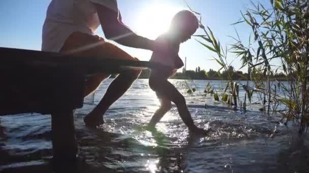 Joven papá sentado en el borde de un embarcadero de madera en el lago y sosteniendo a su hijo en las manos. Niño balanceando los pies en el agua en un día soleado. Familia pasar tiempo juntos al aire libre. Vista lateral Primer plano — Vídeos de Stock