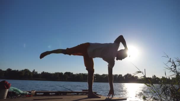 Silhouette di giovane uomo in piedi a posa yoga su un molo di legno al lago. Un ragazzo sportivo che si allena alla natura. Atleta che fa esercizio all'aperto. Luce solare sullo sfondo. Stile di vita sano e attivo. Da vicino. — Video Stock