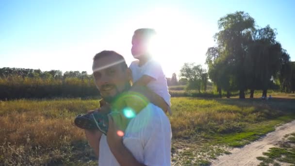Счастливая семья идет по лугу, а папа машет рукой в камеру. Маленький мальчик с светлыми волосами сидит на плечах своего папочки. Молодой отец и ребенок проводят время вместе на природе. Закрыть — стоковое видео