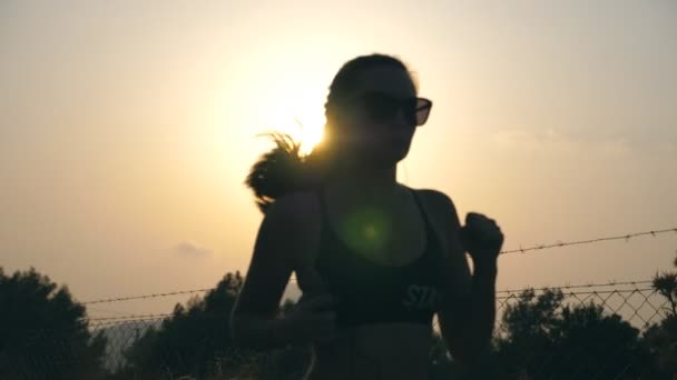 Silhuett av sportig flicka jogging på landet väg vid solnedgången och lyssna musik. Ung kvinna i solglasögon springer utomhus vid solnedgången. En hälsosam aktiv livsstil. Långsam rörelse Närbild — Stockvideo