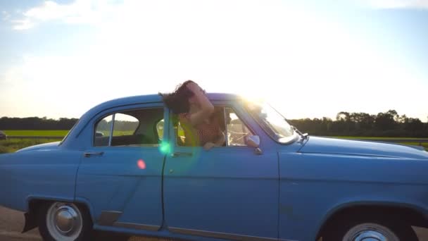 Lycklig flicka i solglasögon lutar ut retro bilfönstret och njuta av resan. Ung kvinna tittar ut fönstret flytta gamla auto. Solen blossa på bakgrunden resor och frihet koncept. Slow motion närbild — Stockvideo