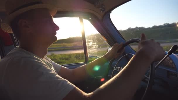Młody człowiek w kapeluszu trzymający się za ręce na kierownicy i jeżdżący retro samochodem z rozbłyskiem słońca w tle. Biały facet podróżujący zabytkowym autem na wsi. Widok z boku Powolny ruch Zamknij — Wideo stockowe