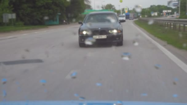 Vue à travers le verre arrière de la voiture en mouvement à l'automobile noire qui conduit sur la route et les phares clignotants le jour couvert. Gouttes de pluie sur la vitre du véhicule après la pluie. POV Mouvement lent — Video