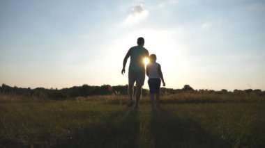 Baba ve oğul sahada günbatımında yürüyüş ve birbirlerinin ellerini tutarak silüeti. Mutlu aile birlikte vakit açık. Aktif yaşam tarzı. Ağır çekim arka geri görüntülemek