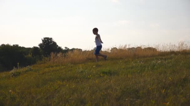 Дитина біжить на галявині на відкритому повітрі. Щасливий усміхнений хлопчик розважається в природі на літньому лузі. Молодий хлопчик з піднятими руками біжить на зеленій траві на полі в сонячний день. Повільний рух крупним планом — стокове відео
