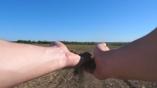 Мужские руки фермера держат груду сухой земли и выливают ее обратно через пальцы на поле в солнечный день. Концепция сельскохозяйственного бизнеса. Синее небо на заднем плане. POV Slow Motion Close up — стоковое видео
