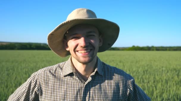 Zblízka šťastný mladý zemědělec v klobouku dívá a usmívá se do kamery na pozadí zelených pšeničné pole. Portrét pohledný muž v košili stojícího na louce na slunečný den — Stock video