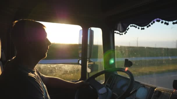 Ο άνθρωπος οδήγηση ενός φορτηγού και να παρακολουθούν με μεγάλη προσοχή στο δρόμο. Καυκάσιο τύπο είναι ιππασία μέσα στο τοπίο στο ηλιοβασίλεμα φόντο. Προφίλ του οδηγός φορτηγού μέσα στην καμπίνα. Εσωτερικη πλευρά άποψη αργή κίνηση — Αρχείο Βίντεο