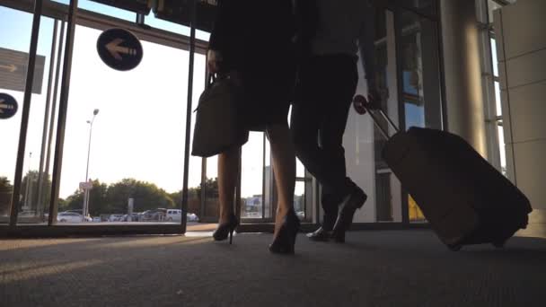 Businessman with his female colleague leaving terminal through glass doors and roll suitcase on wheels. Business man and woman going from the airport to city street with their luggage. Slow motion — Stock Video