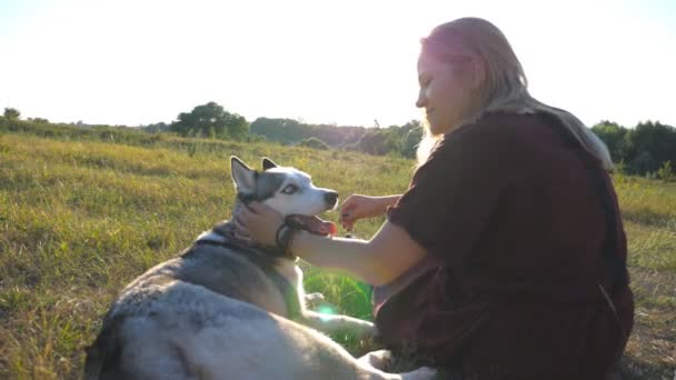 Perfil de chica feliz con el pelo rubio sentado en la hierba verde en el campo y acariciar a su perro husky siberiano en el día de verano. Mujer joven pasando tiempo con su mascota en el prado. Primer plano: cámara lenta — Vídeos de Stock