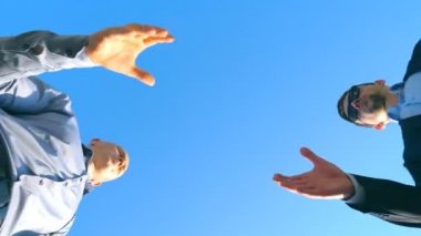 Toplantı ve açık el sıkışan iki başarılı yakışıklı işadamları düşük açılı görünümü. Genç meslektaşım ortağı için mavi gökyüzü arka plan üzerinde siyah bir evrak çantası geçen resmi elbiseli. Yakın çekim