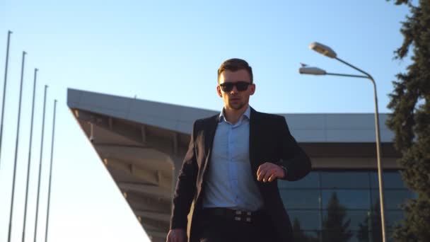 Young businessman in a formal black suit looking at his watch and quickly going with his luggage. Handsome man in sunglasses being late for the meeting or work after flight. Close up Low angle view — Stock Video