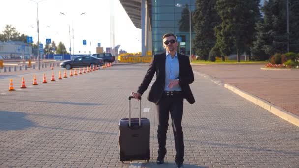 Gelukkige jonge zakenman in een formeel zwart pak grappig dansen en zingen terwijl gaan met bagage op straat na de vlucht. Vrolijke knappe man viert zijn prestatie en succes. Sluiten. — Stockvideo