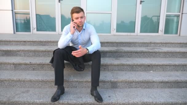 Dolly shot of desperate young manager talking on smart phone and sitting on stairs near office building. Upset sad businessman got a very bad news on cell phone and crying outdoor. Close up — Stock Video