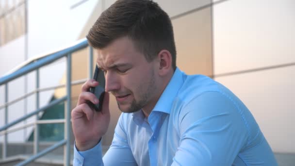 Profili akıllı telefonda konuşurken ve Ofis Binası yakınındaki merdivenlerde oturan çaresiz genç iş adamı. Çok kötü bir haber cep telefonu ve ağlamaya açık Yöneticisi var üzgün üzgün kapatın. Yan görünüm — Stok video