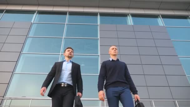 Visão de baixo ângulo de dois jovens empresários felizes com pastas engraçadas dançando enquanto caminham depois de negócios bem sucedidos ao ar livre. Colegas alegres celebrando sua realização perto do centro de escritórios — Vídeo de Stock