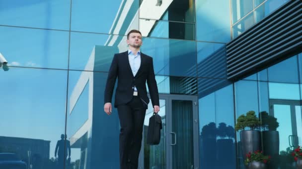 Joven hombre de negocios con un maletín caminando por la calle de la ciudad cerca del moderno edificio de oficinas. El tipo de confianza en traje está de camino al trabajo. Retrato del hombre de negocios viajando al trabajo. Cámara lenta Primer plano — Vídeos de Stock