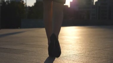 Yüksek topuklu kadın bacakları arka planda güneş ışığıyla şehir caddesinde yürüyor. Şehirdeki yüksek topuklu ayakkabılı genç iş kadınının ayakları. İşe giden bir kız. Yavaş çekim