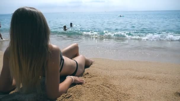 Νεαρή κοπέλα με μπικίνι ξαπλωμένη στην παραλία και κάνει ηλιοθεραπεία. Όμορφη αγνώριστη γυναίκα χαλαρώνοντας στην ακτή του ωκεανού κατά τη διάρκεια των καλοκαιρινών διακοπών ταξίδια. Έννοια της ανάπαυσης στην ακτογραμμή θέρετρο. Οπίσθια όψη — Αρχείο Βίντεο